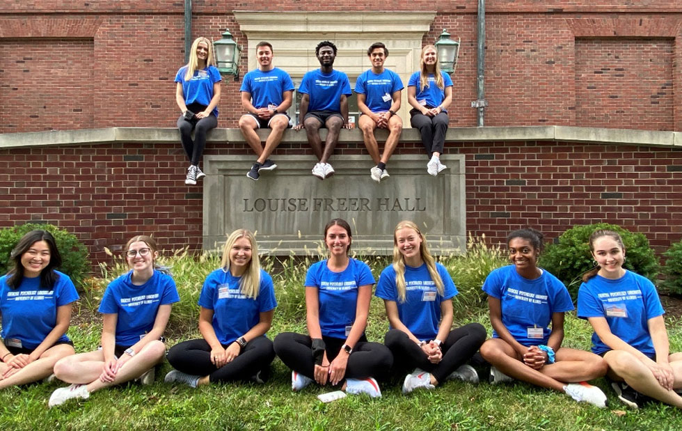 12 undergraduates in matching blue t-shirts sitting in front of Freer Hall